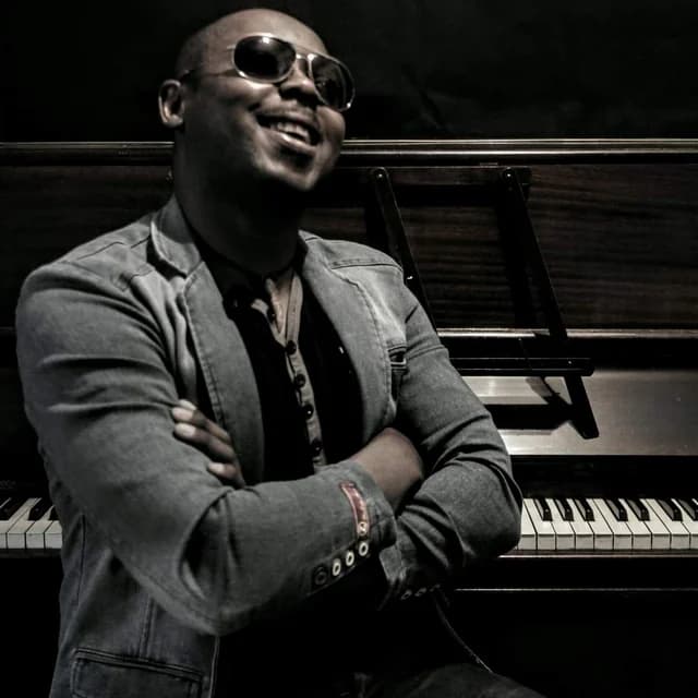 Serge Tebu laughing in sunglasses, arms folded, in front of a piano
