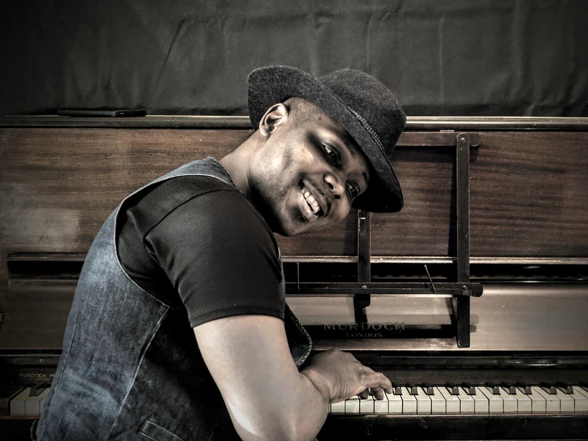 Serge Tebu in a hat at an upright piano, smiling over his shoulder