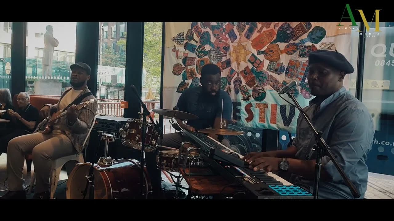 A trio playing outdoors at a festival: guitar, drums and keys in front of a patterned banner
