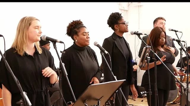 Five singers at microphones in a bright rehearsal room, a band behind them
