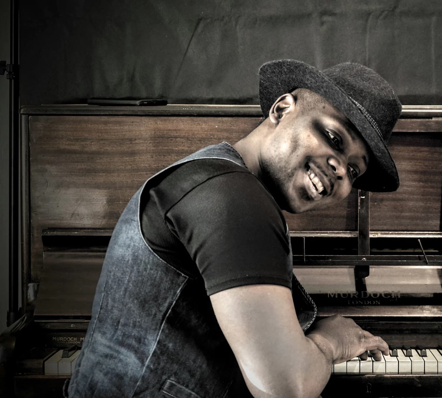Serge Tebu in a hat at an upright piano, smiling over his shoulder, one hand on the keys