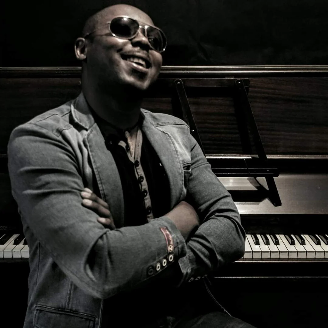 Serge Tebu laughing in sunglasses, arms folded, in front of an upright piano